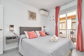 Habitación de hotel con cama, mesitas de noche, aire acondicionado de pared, ventana con cortinas y balcón con una silla.