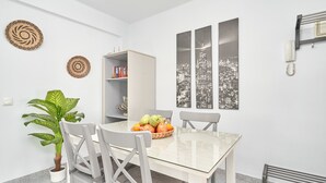Un coin repas avec une table en verre, des chaises blanches, une plante en pot, un panier de fruits et une œuvre d’art représentant un paysage urbain accrochée au mur.