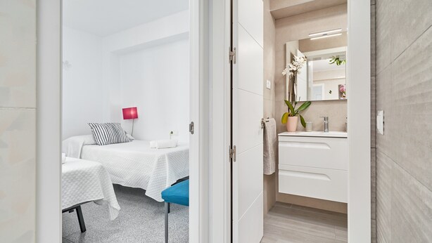 Une chambre d’hôtel moderne comprenant un lit, des tables de chevet, un petit bureau, une chaise, une lampe rouge et une salle de bain avec un miroir, un lavabo et une plante en pot.