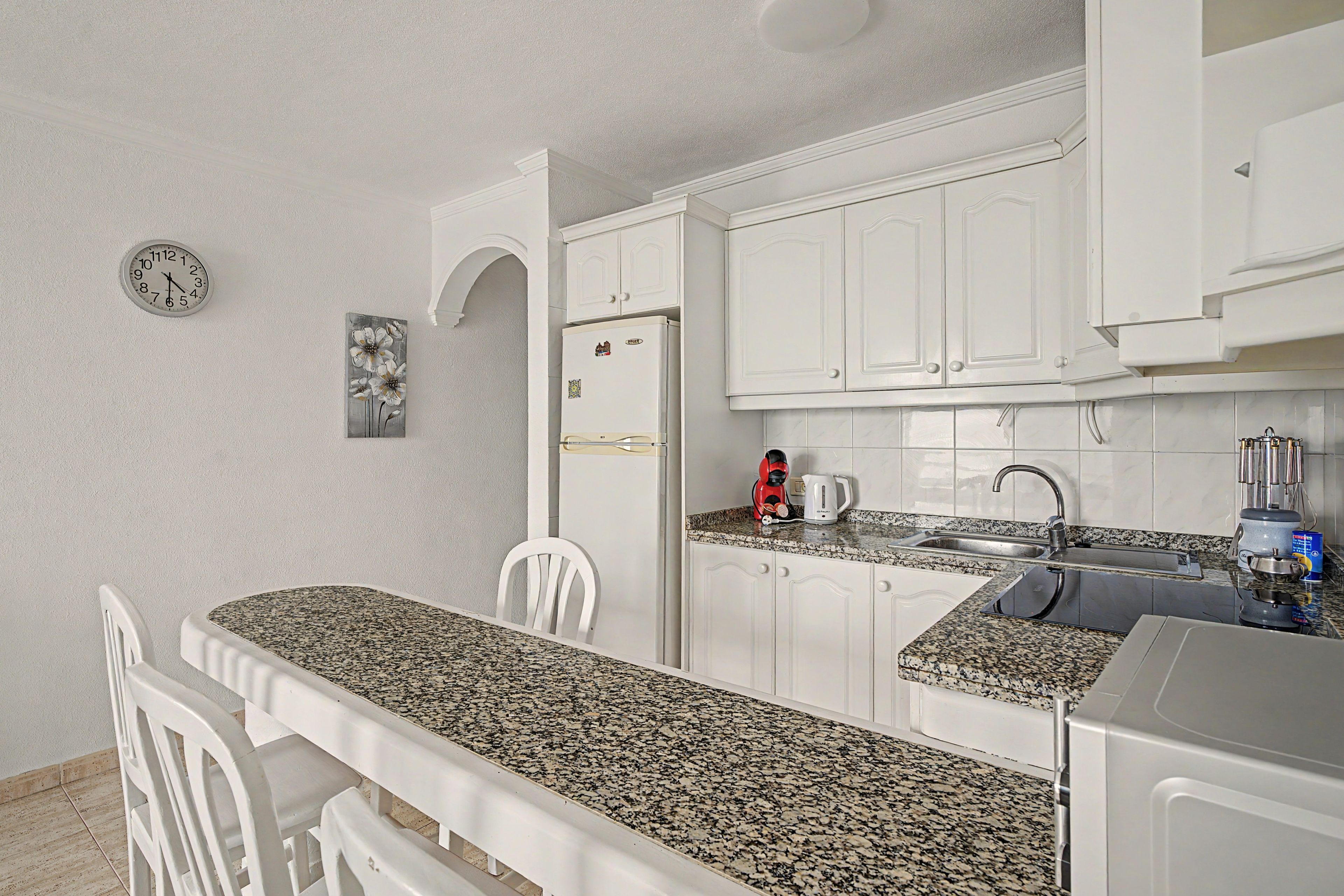 A kitchen with white cabinets, a granite countertop, a clock, and a refrigerator.