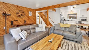 Een moderne woonkamer met twee grijze banken, een houten salontafel en een trap naar een keuken met witte kasten en een magnetron.