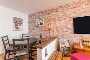 A room with a brick wall, a round table with chairs, a bar area with a jukebox, and a flat-screen TV mounted on the wall.