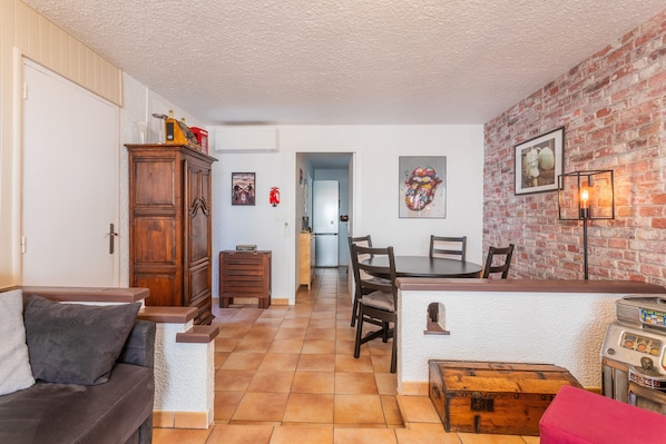 A living room with a sofa, a wooden cabinet, a dining table with chairs, a kitchen in the background, and a brick wall.
