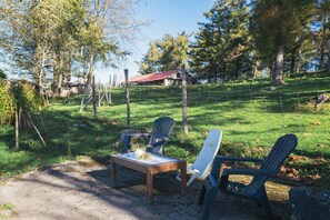 Uma área de estar ao ar livre rústica, com mesa de madeira e cadeiras Adirondack, cercada por um pasto e árvores.