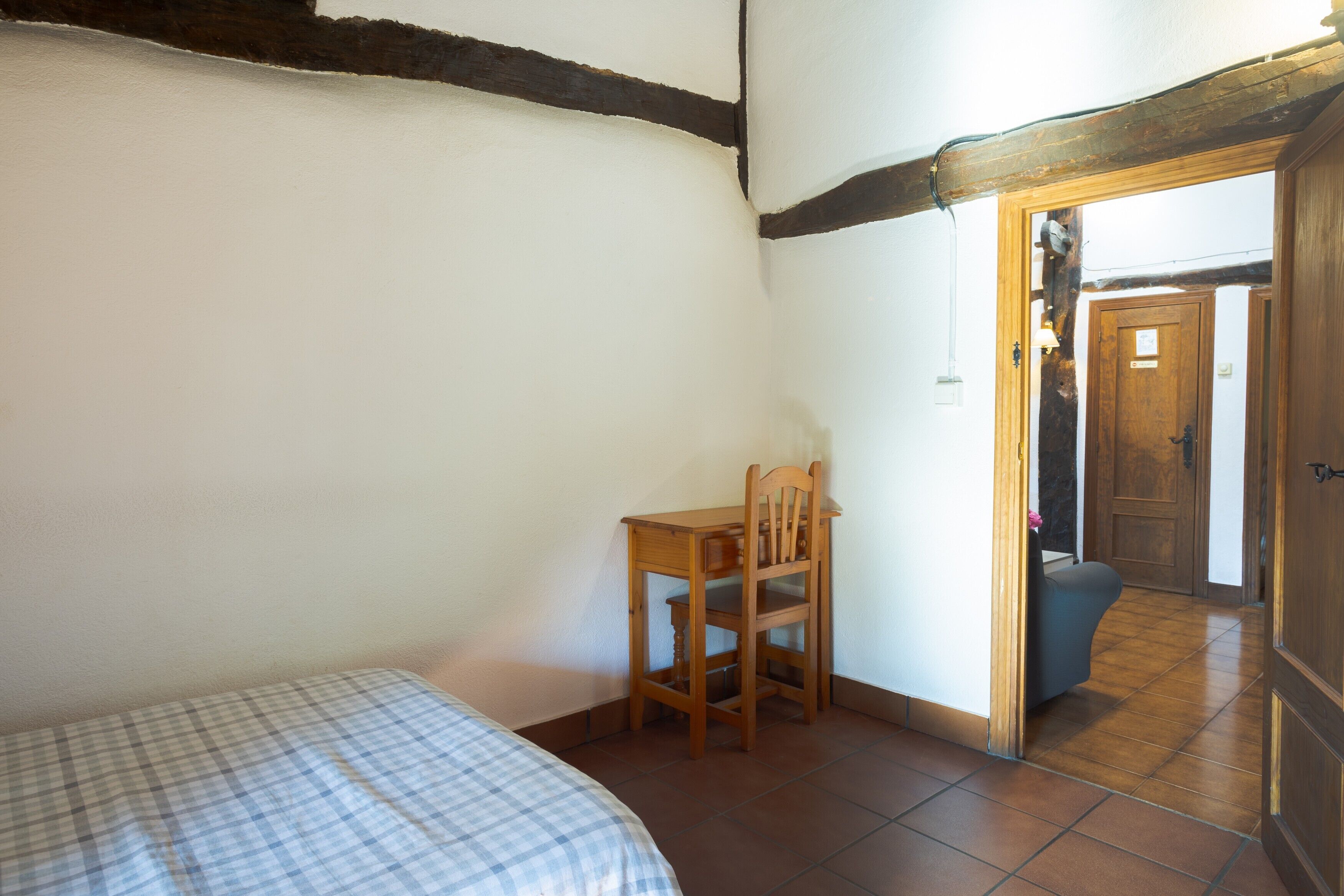 Un dormitorio con cama, una silla de madera, una mesita y una puerta que da a otra habitación.