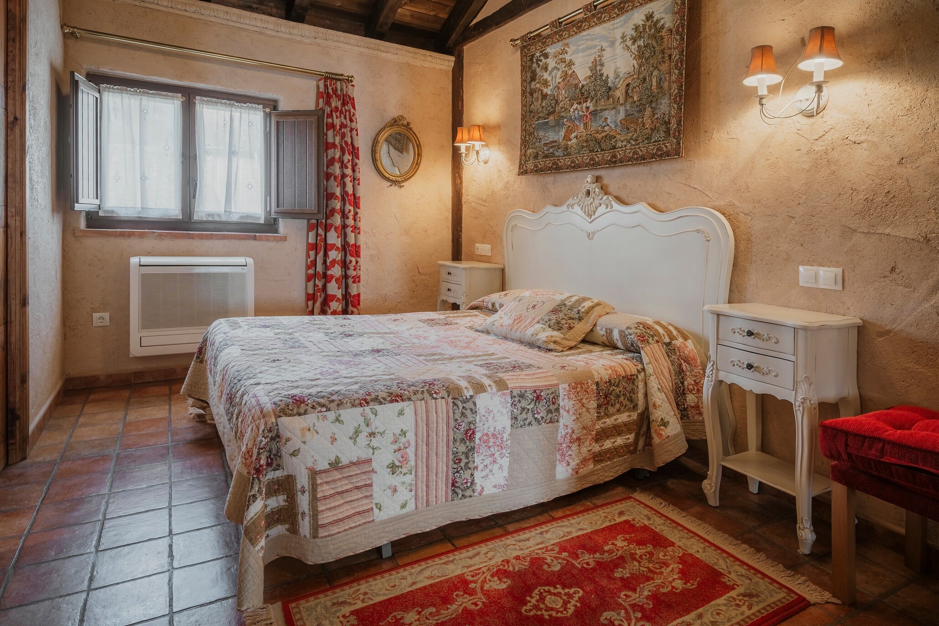 A bedroom with a bed, a nightstand, a red chair, a window with curtains, a wall-mounted lamp, and a painting.