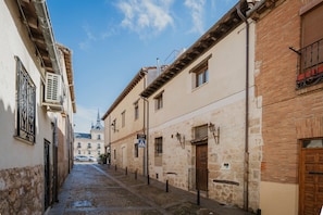 Una estrecha calle empedrada flanqueada por edificios de fachadas de piedra, una aguja de iglesia a lo lejos, y un cielo azul despejado.