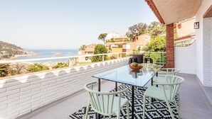Ein Balkon mit Glastisch und Stühlen, einem gemusterten Teppich und Blick auf das Meer sowie anliegende Gebäude am Hang.