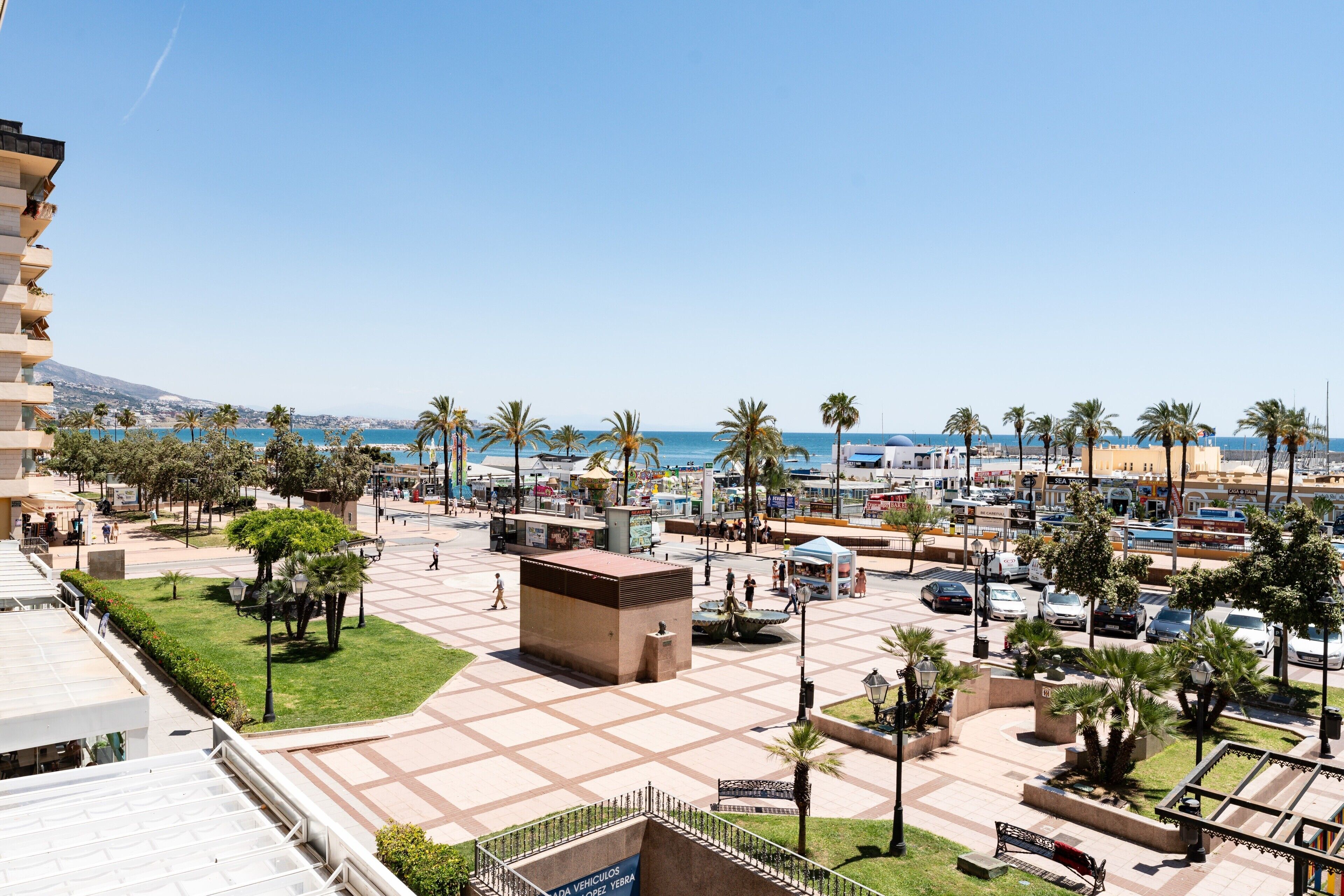 Una piazza con palme, una fontana e vista sul mare.