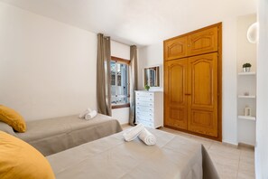 Une chambre avec deux lits, une commode, une armoire, une fenêtre avec des rideaux et une petite étagère avec une plante.