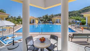 A poolside area with lounge chairs, a table with fruit, and a view of a swimming pool and surrounding buildings. - Holiday Home 'Dioni' with Mountain View, Wi-Fi and Air Conditioning (Lefkada)