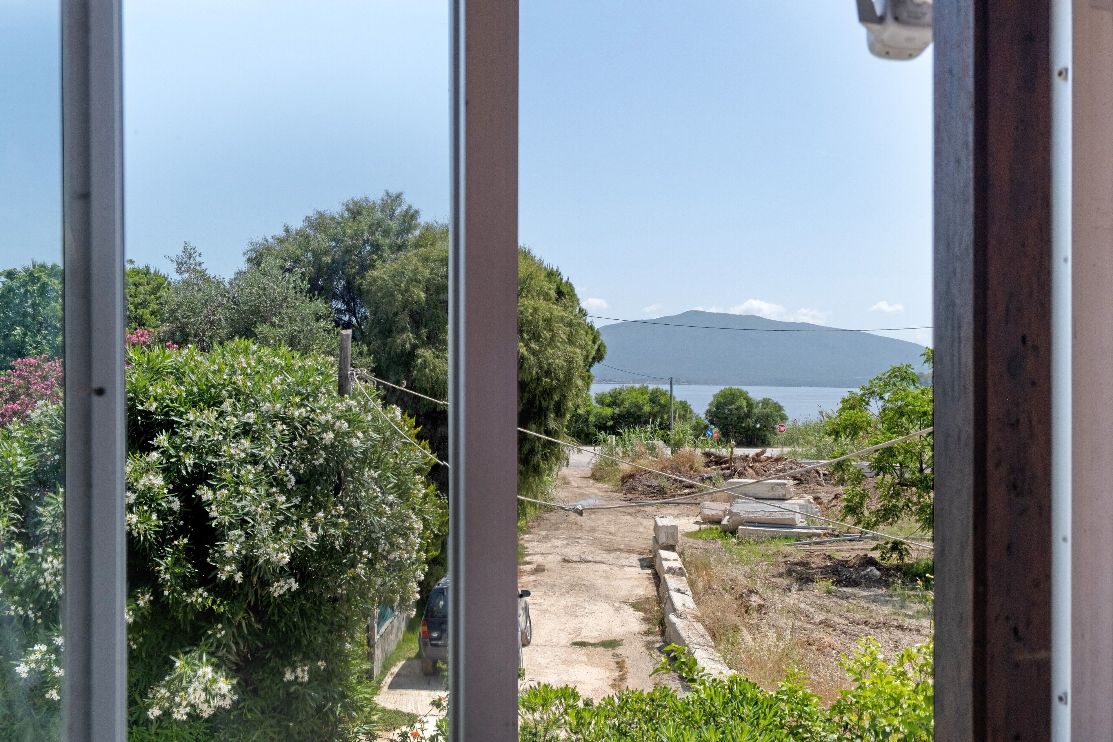 Une vue à travers une fenêtre sur un chantier de construction, avec un chemin de terre, de la végétation et une montagne au loin.