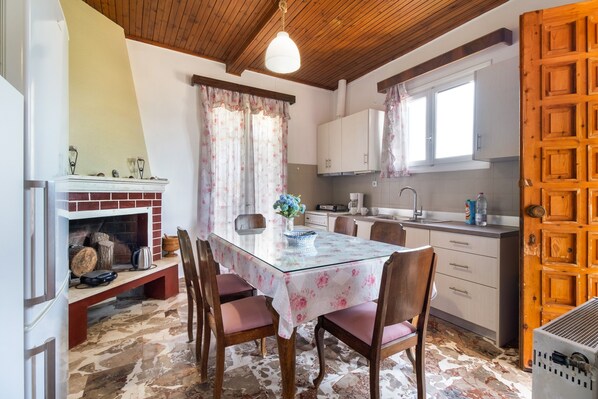 A dining area with a glass table, wooden chairs, a fireplace, and a kitchen in the background.