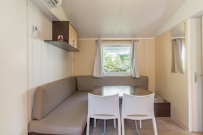 Een kleine kamer met een bank, een tafeltje, twee stoelen, een raam met gordijnen, een plank met een apparaat en een deur.