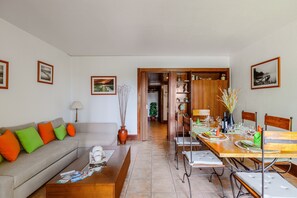 A living room with a sofa, coffee table, dining table set for four, and a hallway leading to another room.
