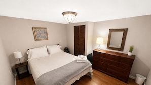 Ein Schlafzimmer mit einem Bett, Nachttischen, einer Kommode, einem Spiegel, einer Deckenleuchte und einem gerahmten Bild an der Wand.