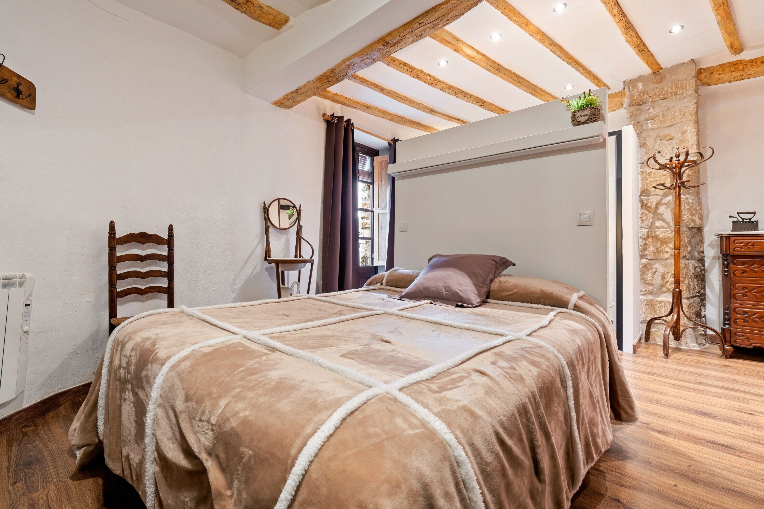 A bedroom with a large bed, wooden beams, a stone wall, and a coat rack.