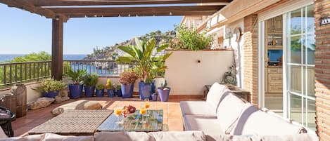 A covered patio with wicker furniture, a glass table with fruit, and potted plants.