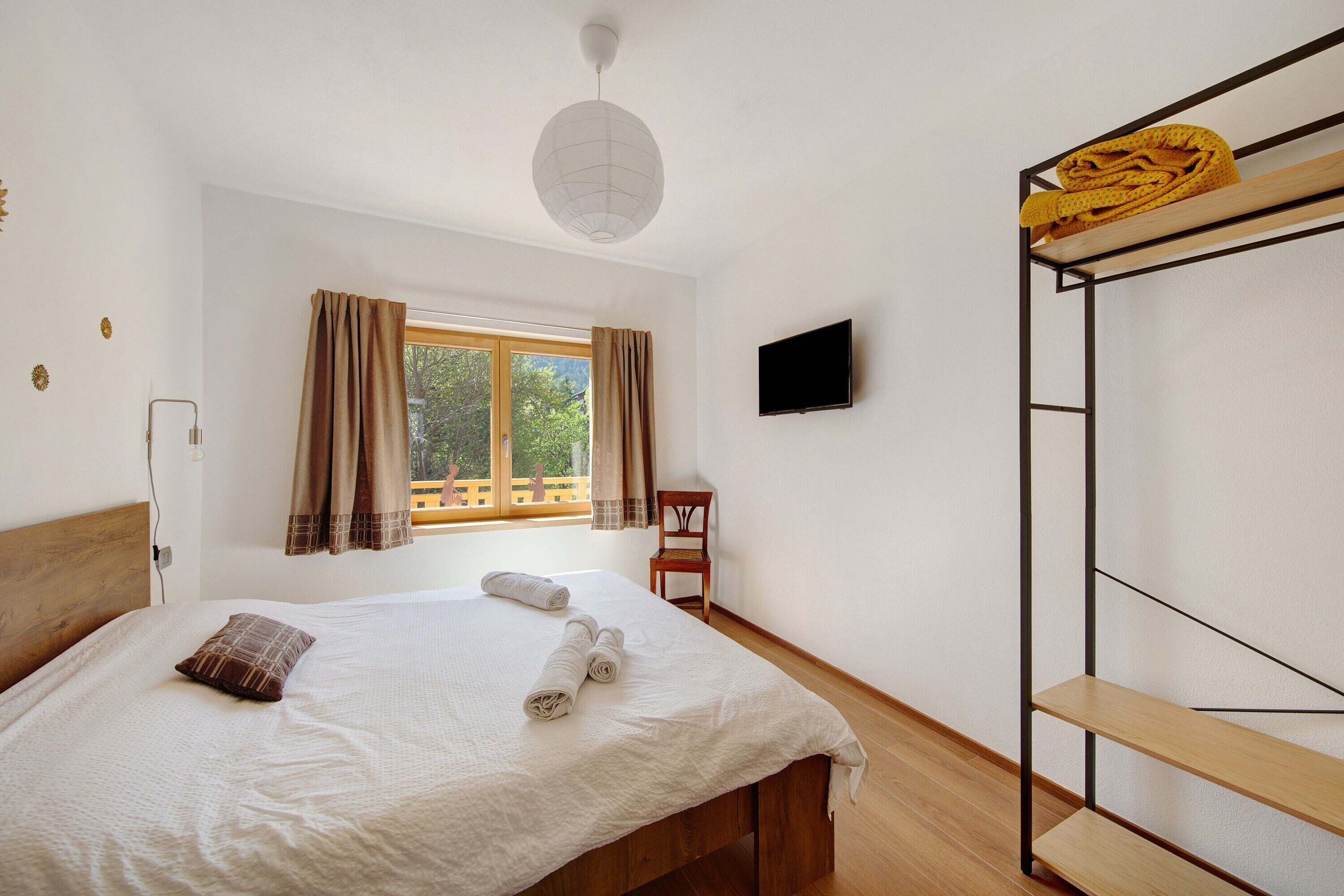 Un dormitorio con una cama, una silla, un televisor, un ventanal con cortinas y una litera.