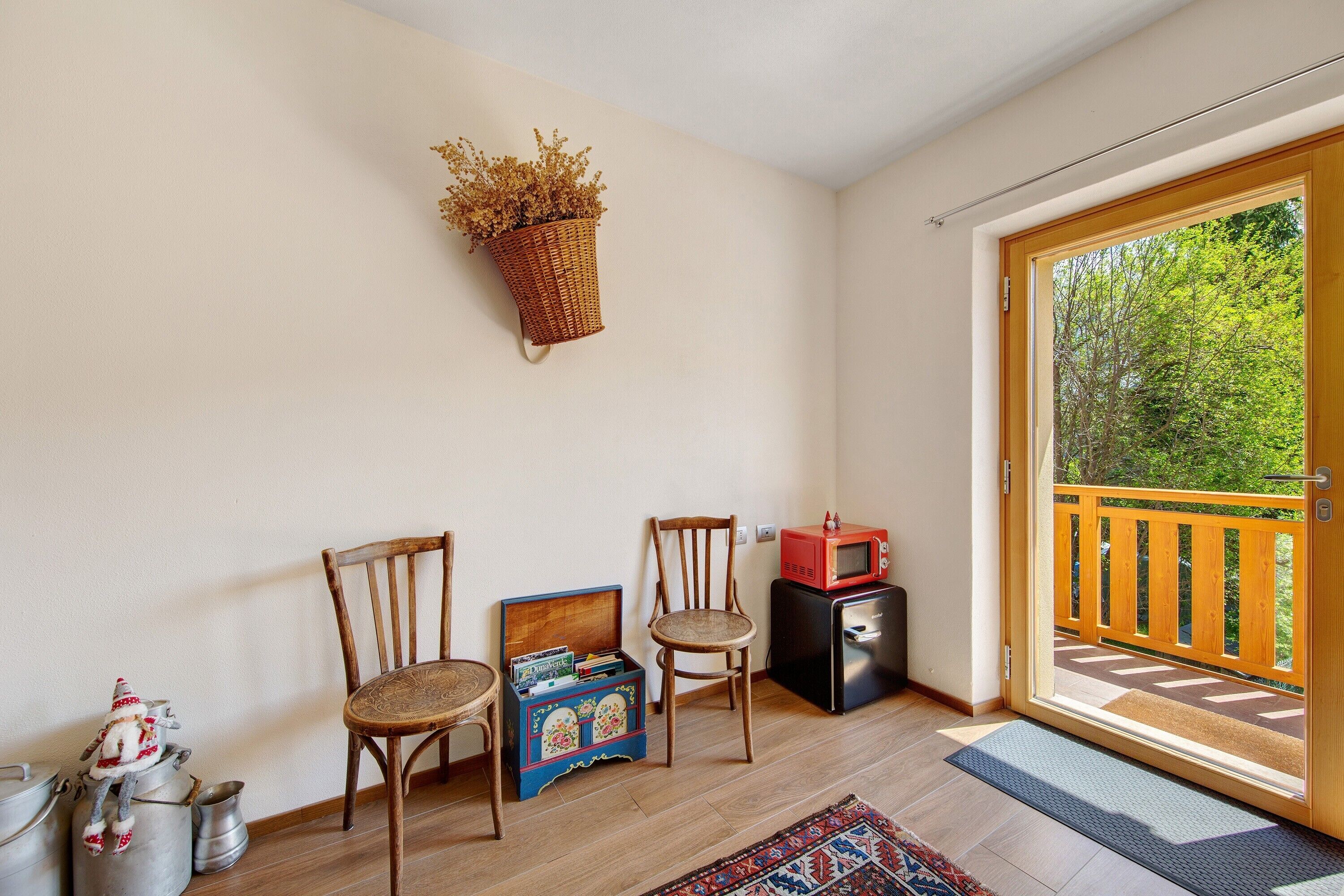 Una habitación con dos sillas de madera, una cesta de mimbre en la pared, un electrodoméstico negro pequeño y una puerta de madera que da a un balcón.