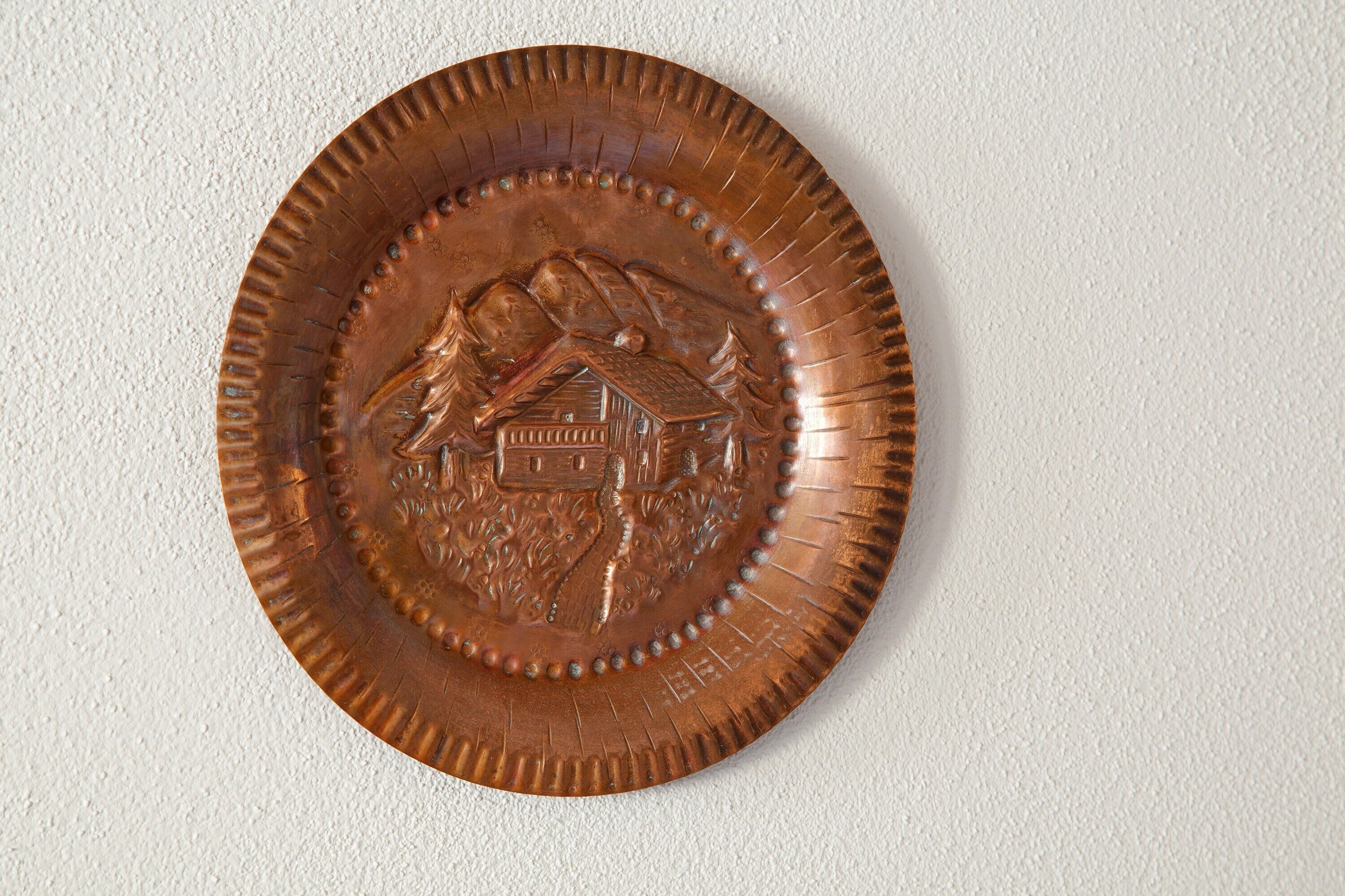Une plaque circulaire en cuivre ornée d’un motif représentant une maison et une montagne.