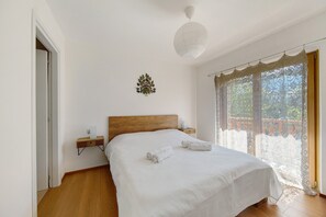 A bedroom with a wooden headboard, a bedside table with a lamp, a large bed with white linens, and a view through a window with lace curtains. - Room 4 – Nene Maria with Terrace and Shared Garden (Pieve di Cadore)