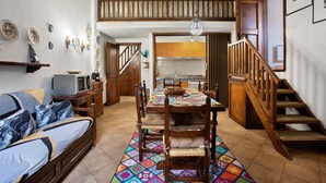 Un coin repas avec une table en bois, des chaises et un tapis coloré. Un escalier avec des rampes en bois mène à un niveau supérieur.