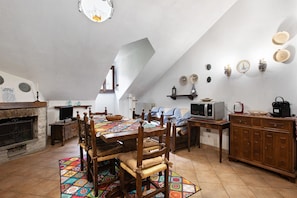 Comedor con mesa de madera, sillas, alfombra estampada, chimenea y zona de cocina con electrodomésticos.