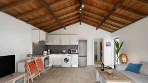Eine moderne Küche mit weißen Schränken, einer Waschmaschine und einer Mikrowelle. Im Wohnbereich befindet sich ein Holztisch und eine Couch.