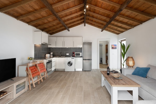 A modern kitchen with white cabinets, a washing machine, and a microwave. There is a wooden dining table and a sofa in the living area.