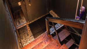 Eine Holztreppe mit einer kleinen Ablage, auf der zwei Blumentöpfe stehen, führt zu einer dunklen Tür.