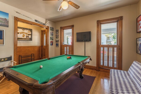 A pool table in a room with wooden floors, a ceiling fan, and a television on the wall.