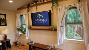 A cozy living room with a TV displaying "JOE'S RIDGE RETREAT" on the wall, a wooden bench, a sofa, and a small table with books. - Jungle Boogie in Mountains (Cool Ridge)