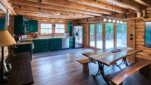 L’intérieur d’un chalet rustique, avec des poutres en bois, une cuisine aux armoires vertes, une table à manger et une grande fenêtre donnant sur une zone boisée.