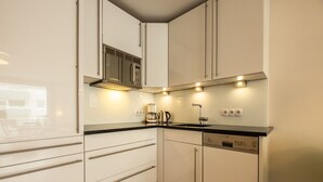 A compact kitchen with white cabinets, a black countertop, and built-in appliances including a coffee maker and a dishwasher. - Malin Haus Eydum - 2 room apartment in Haus Eidum. (Sylt)
