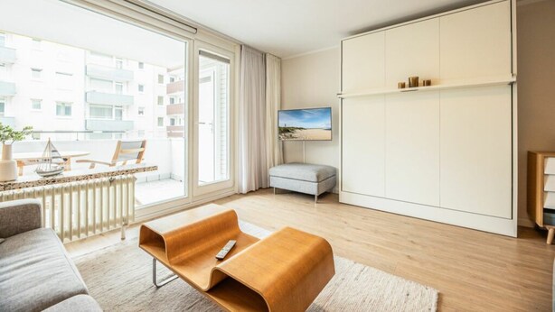 A modern living room with a sofa, a coffee table, a side table, a large window, a sliding door, a painting, and a wardrobe. - Malin Haus Eydum - 2 room apartment in Haus Eidum. (Sylt)
