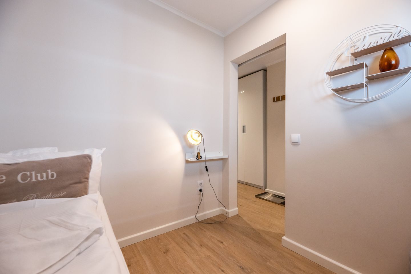 A hotel room with a bed, a bedside table with a lamp, a wall-mounted shelf with a vase, and a door leading to another room.