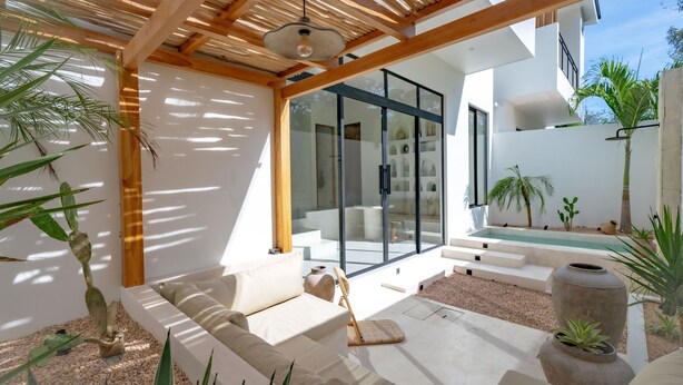 A modern outdoor living area with a wooden pergola, a white sofa, and a glass door leading to a pool. - Casa Terra Uluwatu (Pecatu)