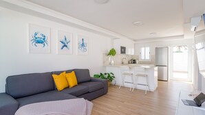Un soggiorno moderno con un divano grigio, cuscini gialli e una coperta beige. Si intravede sullo sfondo una cucina con elettrodomestici bianchi e una zona pranzo con sedie bianche.