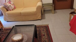 A beige sofa with a floral pillow, a glass-top coffee table with a decorative item, a patterned rug, and a small stool. - LOCRI Epizephiri - Apartment 3 pl - Very close to the sea  (Locri)