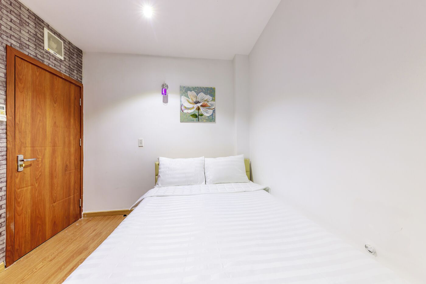 A bedroom with a wooden door, a bed with white bedding, a wall-mounted light, and a floral painting.