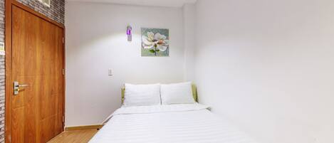 A bedroom with a wooden door, a bed with white bedding, a wall-mounted light, and a floral painting.