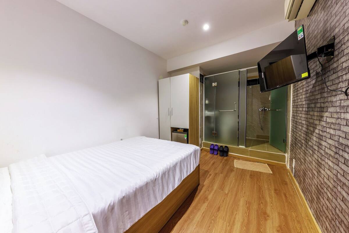 Habitación de hotel con cama, televisor montado en la pared, puerta corredera que da a una zona de ducha y suelo de madera.