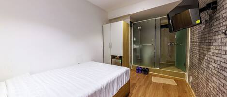A hotel room with a bed, a TV mounted on the wall, a sliding door leading to a shower area, and a wooden floor.