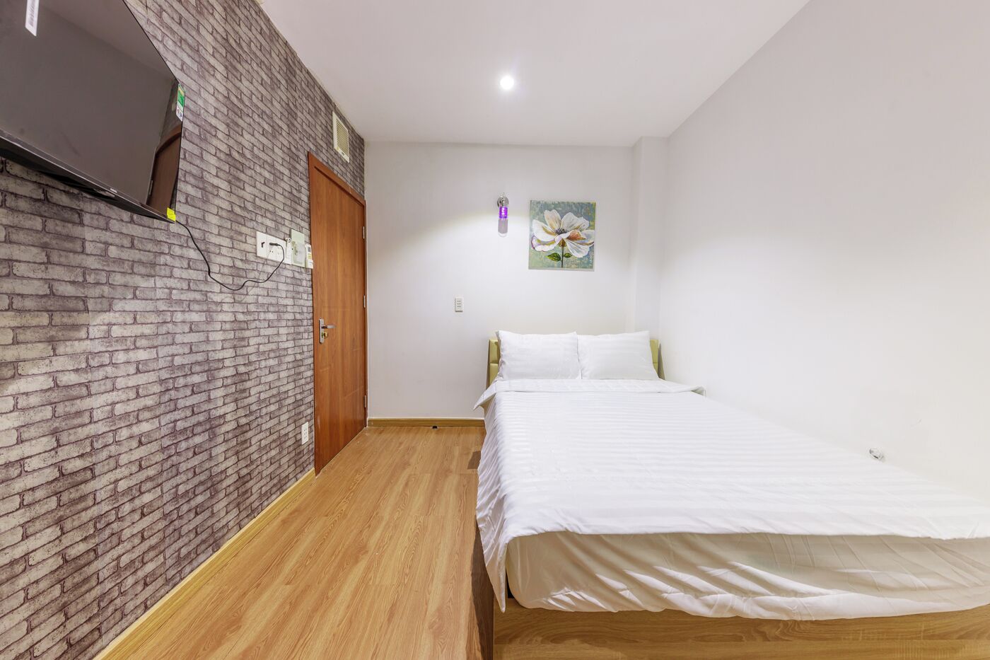 Une chambre avec un mur en briques, un plancher en bois, un lit avec une literie blanche, une porte en bois et un tableau accroché au mur.