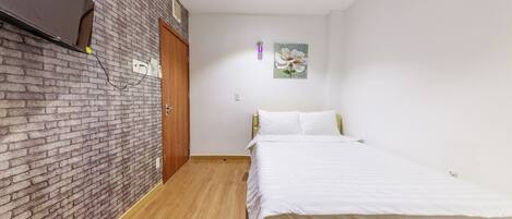 Une chambre avec un mur en briques, un plancher en bois, un lit avec une literie blanche, une porte en bois et un tableau accroché au mur.
