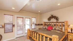 A bedroom with a wooden bed frame, a headboard with a decorative wreath, and a red pillow. There are two doors with blinds, a carpeted floor, and a nightstand with a lamp. - 3-bedroom condo in fabulous Eden, skiing, biking, hiking, and water sports (Eden)