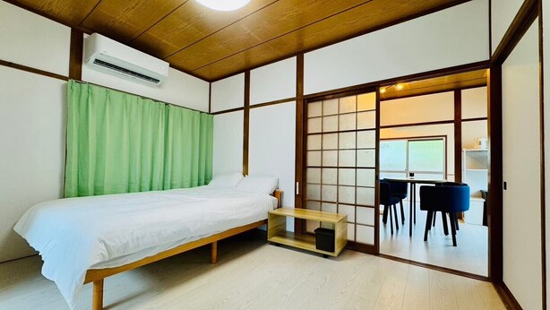 Una habitación tradicional japonesa con una cama de madera, un futón, una puerta corredera que da a un comedor y un equipo de aire acondicionado.