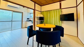 Una habitación con una mesa redonda, dos sillas, un televisor montado en la pared y una cocina visible a través de un vidrio.