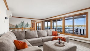Un salon spacieux avec un grand canapé d’angle, un petit pouf et une vue sur un lac grâce à de grandes fenêtres.